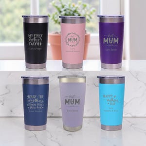 Op de afbeelding: Een verzameling geïsoleerde bekers in diverse kleuren, waaronder zwart, roze, paars, marineblauw, lavendel en turquoise. Elke beker heeft een zilveren rand en deksel, met gepersonaliseerde tekst zoals "My First Mother's Day" en "Best Mum Ever".