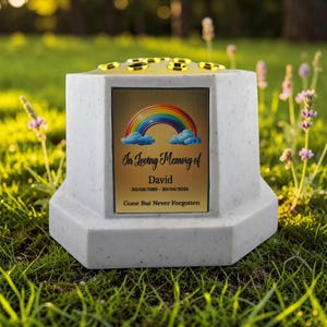Puede incluir: Un jarrón conmemorativo blanco con una placa dorada con un arcoíris y nubes. La placa dice "In Loving Memory of David" con fechas y "Gone But Never Forgotten". La parte superior tiene detalles dorados.