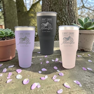 Op de afbeelding: Drie geïsoleerde bekers in lavendel, houtskool en roze, elk met een zilveren deksel. De bekers hebben de tekst "I DON'T SNORE I DREAM I'M A Motorcycle Love Sam" en een motorfietsafbeelding. De bekers staan op een houten oppervlak.