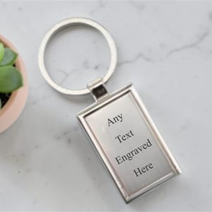 May include: Silver rectangular keychain with the text "Any Text Engraved Here." The keychain has a silver ring and a rectangular plate. A small potted succulent plant is in the upper left corner.