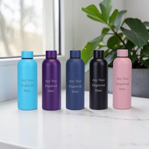 May include: Five water bottles in shades of blue, purple, navy, black, and pink, each with a matching cap. The bottles have the text "Any Text Engraved Here" printed on them. They are arranged on a white surface.