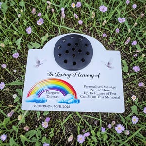 Puede incluir: Placa conmemorativa blanca con un portaflores negro, un arcoíris e ilustraciones de palomas. El texto incluye "In Loving Memory of", un nombre, fechas y espacio para un mensaje personalizado. En un lecho de hierba y flores.