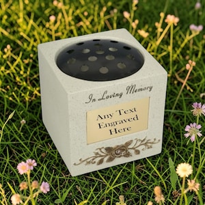 Op de afbeelding: Een vierkante, lichtgekleurde herdenkingsvaas met een zwarte bloemenkikkerinzet. De vaas heeft een goudkleurige plaat met de tekst "Any Text Engraved Here" en de zin "In Loving Memory". Onder de plaat staat een bloemenontwerp.