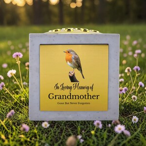 May include: A square memorial urn with a gold-colored center panel featuring a robin perched on a branch. The text reads "In Loving Memory of Grandmother, Gone But Never Forgotten." The urn is set in a grassy area with flowers.