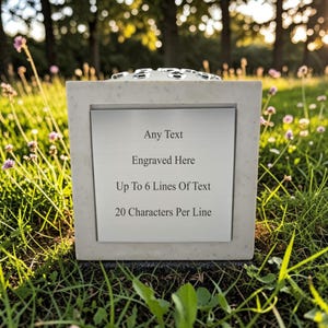 Puede incluir: Una urna conmemorativa cuadrada de color claro con una parte superior decorativa, colocada en una zona cubierta de hierba. La urna presenta una placa plateada con el texto: "Any Text, Engraved Here, Up To 6 Lines Of Text, 20 Characters Per Line."