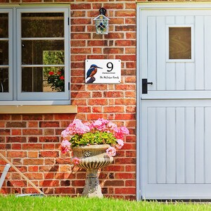 May include: A white metal house number sign with a blue kingfisher bird illustration. The sign reads "9,  Chimneys Street, The McElynn Family".