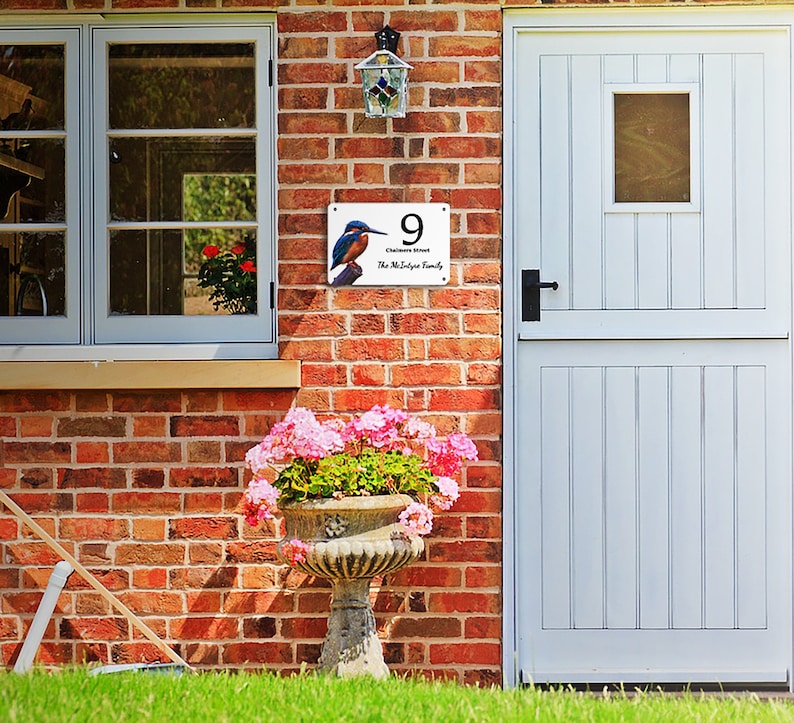 May include: A white metal house number sign with a blue kingfisher bird illustration. The sign reads "9,  Chimneys Street, The McElynn Family".
