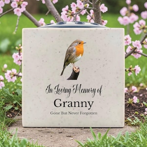 May include: A square, speckled, cream-colored memorial vase with a robin perched on a branch. The text "In Loving Memory of Granny, Gone But Never Forgotten" is printed in black script. The vase has a dark blue top.