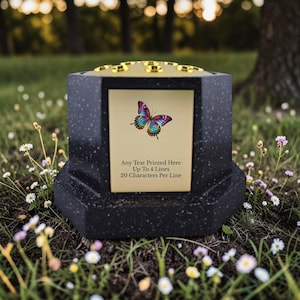 May include: A black, hexagonal memorial urn with a gold-coloured top and a gold plaque featuring a colourful butterfly illustration. The plaque includes the text: "Any Text Printed Here Up To 4 Lines 20 Characters Per Line."
