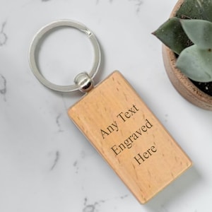 May include: A rectangular wooden keychain with a silver key ring. The keychain has the text "Any Text Engraved Here" engraved on it. A small succulent plant in a brown pot is in the background.