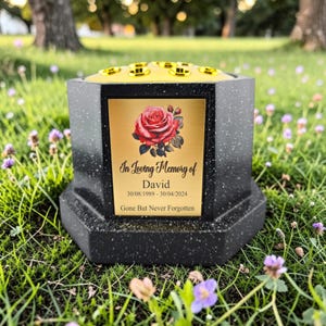 Puede incluir: Un jarrón conmemorativo negro y dorado con una placa dorada con una rosa roja y la inscripción "In Loving Memory of David 30/08/1989-30/04/2024 Gone But Never Forgotten." El jarrón tiene forma hexagonal y está sobre hierba.