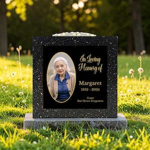 May include: A black granite headstone with a photo of Margaret, 1952-2024, in a gold oval frame. The headstone has the inscription "In Loving Memory of Margaret" and "Gone But Never Forgotten" in gold script.