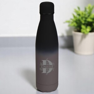 May include: A matte black water bottle with a gradient finish transitioning to a dark grey base. The bottle features a centred, engraved design consisting of a large letter D with the name David written horizontally across the centre.