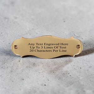 May include: A gold-coloured metal plaque with a decorative curved shape and two small circular holes on the ends for mounting. The centre features the engraved text: Any Text Engraved Here, Up To 3 Lines Of Text, 20 Characters Per Line.