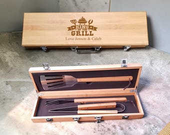 Puede incluir: Un juego de utensilios de barbacoa de bambú en un maletín de madera rectangular. La tapa tiene grabado el texto KING OF THE GRILL y Love Jenson & Caleb. En el interior, una espátula, un tenedor y unas pinzas de metal con mangos de bambú.
