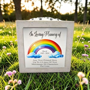 Könnte beinhalten: Quadratische, hellfarbene Gedenkurne mit dekorativer Oberseite. Die Urne hat eine Metallplakette mit dem Text "In Loving Memory of" über einem Regenbogen-Design. Der Text "David 1945-2024" befindet sich darunter. Zusätzlicher Text ist ebenfalls vorhanden.