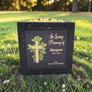 Könnte beinhalten: Eine schwarze Gedenkurne mit einem goldenen Kreuz und der Inschrift "In Loving Memory of Margaret 1952-2024". Die Urne hat eine gesprenkelte Textur und eine goldene Oberseite mit Blumenöffnungen. Die Worte "Gone But Never Forgotten" sind ebenfalls eingeschrieben.