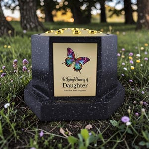 以下が含まれることがあります： カラフルな蝶のイラストが描かれた金色のプレートが付いた黒い記念の骨壺。プレートには「In Loving Memory of Daughter, Gone But Never Forgotten」と書かれています。骨壺は花のある草地の中に置かれています。