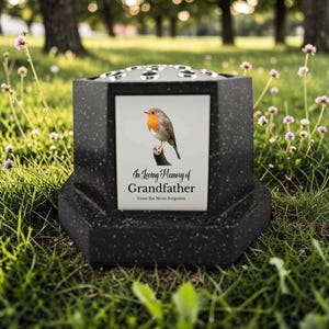 May include: A black memorial urn with a white framed picture of a robin bird perched on a branch. The picture has the text "In Loving Memory of Grandfather, Gone But Never Forgotten". The urn is on a grassy lawn.