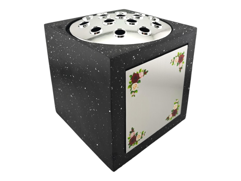 May include: A black speckled cube-shaped vase with a silver top featuring multiple holes for flower stems. A mirrored panel on the front is decorated with floral corner designs in red, pink, and green.
