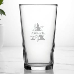 Könnte beinhalten: Ein klares Bierglas mit individuellem Design. Das Design zeigt den Namen "Amanda" in einer Serifenschrift, mit dem Buchstaben "A" über dem Namen und floralen Akzenten. Das Glas ist leer und steht auf einer weißen Oberfläche.