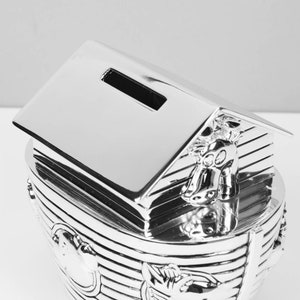 Puede incluir: Una alcanc&iacute;a de plata plateada con forma de arca de No&eacute; con una ranura en el techo para monedas. El arca tiene un dise&ntilde;o detallado con animales en los lados.