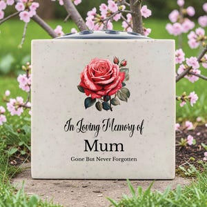 Może przedstawiać: Kwadratowy, kremowy wazon pamiątkowy z czerwonym motywem róży i napisem "In Loving Memory of Mum, Gone But Never Forgotten". Wazon ma ciemną górę z otworami na kwiaty. Różowe kwiaty i zielona trawa w tle.