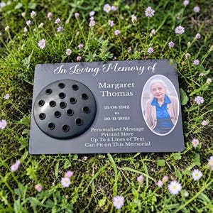 May include: A dark gray memorial plaque with a floral insert and a photo of Margaret Thomas. The plaque includes the dates 21/08/1942 to 30/11/2025 and the text "In Loving Memory of". The plaque is surrounded by green grass and small purple flowers.