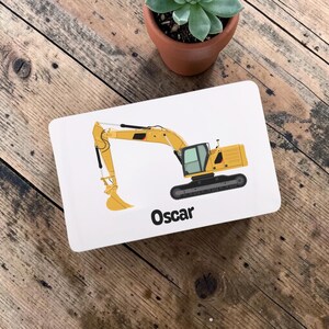 May include: A white rectangular tray featuring a yellow excavator illustration. The excavator has a black track, a detailed arm, and a bucket. The name "Oscar" is printed in black at the bottom of the tray.