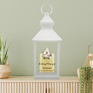 Puede incluir: Linterna blanca con una vela blanca en su interior. La vela tiene un diseño floral y el texto "En memoria cariñosa de mi esposo Se fue pero nunca será olvidado".
