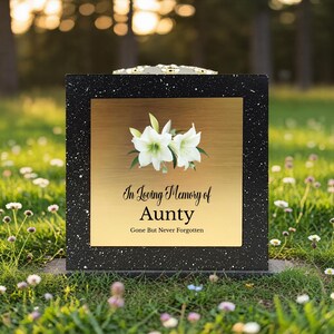 Op de afbeelding: Zwart en goud gedenkplaat met witte bloemen en de tekst "In Loving Memory of Aunty, Gone But Never Forgotten." De plaat staat in een grasveld met bloemen.
