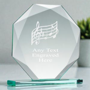 May include: A clear, faceted glass award featuring a musical note design. The text "Any Text Engraved Here" is below the musical notes. The award is set on a rectangular base. A small plant in a white pot is in the background.