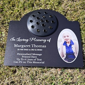 Puede incluir: Placa conmemorativa negra con una foto de una mujer y el texto "In Loving Memory of Margaret Thomas 21/08/1942 to 30/11/2025". La placa tiene un portavelas redondo con agujeros para flores.
