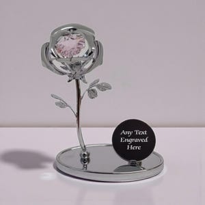 May include: A silver rose sculpture with a pink crystal centre. The rose sits on a round, mirrored base, with a small black disc that reads "Any Text Engraved Here". The sculpture is a decorative item.