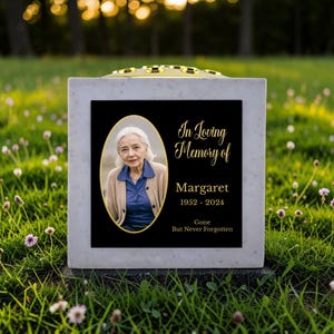 May include: A memorial stone with a black plaque featuring a photo of a woman in an oval frame. Gold text reads "In Loving Memory of Margaret 1952-2024 Gone But Never Forgotten". The stone is set in a grassy area.