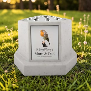 May include: A gray memorial vase with a bird image and the text "In Loving Memory of Mum & Dad". The vase has a hexagonal shape and a top with multiple holes. The vase is sitting on green grass.