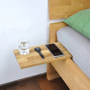 May include: A wooden bedside table with a glass of water, a watch, and a smartphone on top. The table is attached to the side of a bed frame.