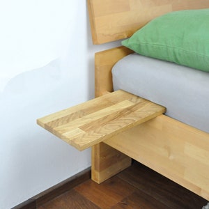 May include: A wooden bed frame with a light green pillow and a small wooden shelf attached to the side. The shelf is made of the same wood as the bed frame and is ideal for holding a book or a glass of water.