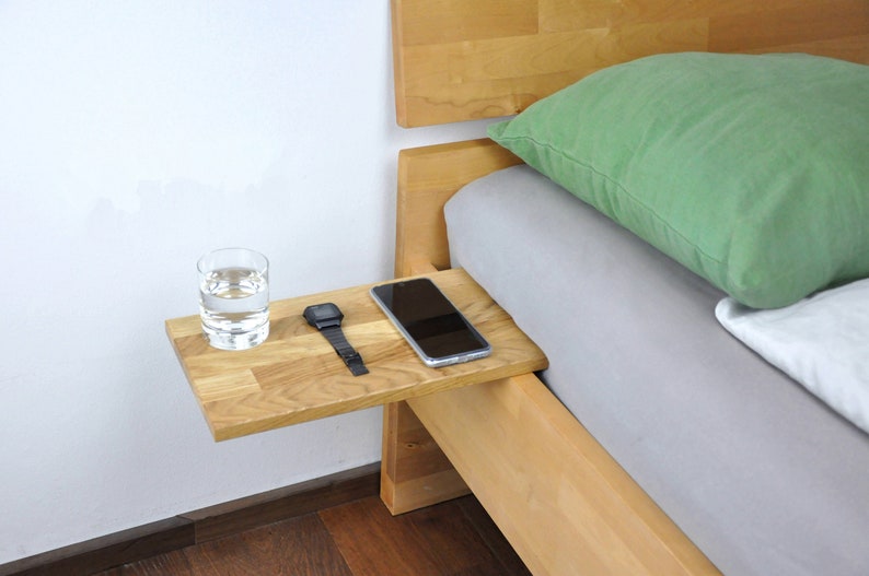 May include: A wooden bedside table with a glass of water, a watch, and a smartphone on top. The table is attached to the side of a bed frame.