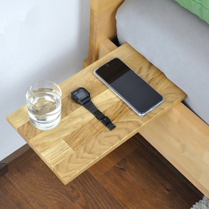 May include: A wooden bedside table with a glass of water, a watch, and a smartphone on top. The table is attached to the side of a bed.