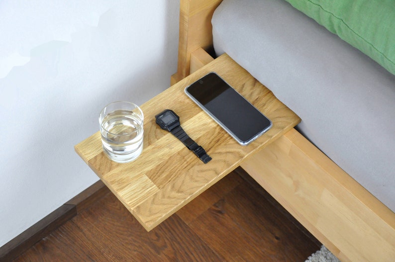 May include: A wooden bedside table with a glass of water, a watch, and a smartphone on top. The table is attached to the side of a bed.