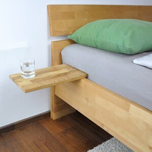 May include: A wooden bedside table attached to a bed frame. The table has a glass of water on it. The bed has a green pillow and a white pillow.