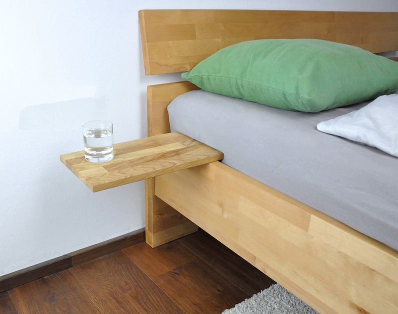 May include: A wooden bedside table attached to a bed frame. The table has a glass of water on it. The bed has a green pillow and a white pillow.
