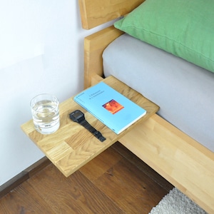 May include: A wooden bedside table with a book, a watch, and a glass of water on top. The table is attached to the side of a bed.
