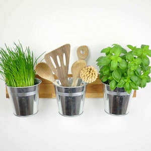 May include: Three silver metal pots with wooden handles are mounted on a wooden shelf. The pots contain green herbs, including chives and basil.