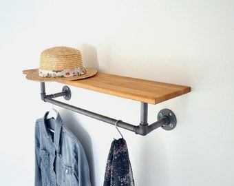 May include: A wooden shelf with a metal pipe rail attached. The shelf is mounted to a white wall. A straw hat, a denim jacket, and a scarf are hanging on the rail.