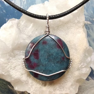 Puede incluir: Un colgante de piedra preciosa azul verdoso y rojo rubí con envoltura de alambre plateado, colgado de un cordón negro.