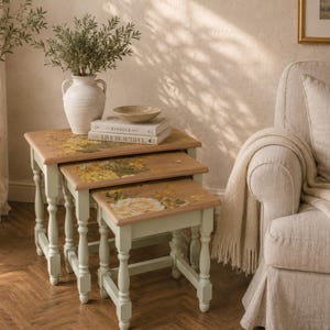 May include: A set of three nesting tables with light green turned legs and natural wood tops featuring floral decals. The tables are stacked in a descending size order, each displaying a printed design of yellow and white roses and green foliage.