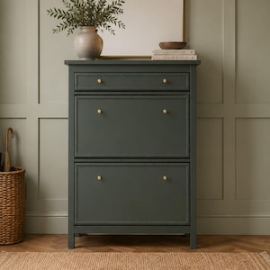 May include: A dark charcoal grey shoe storage cabinet featuring one top drawer and two large tilt-out compartments. The cabinet is accented with small, round, gold-toned knobs on each drawer and compartment front, providing a clean, functional design.