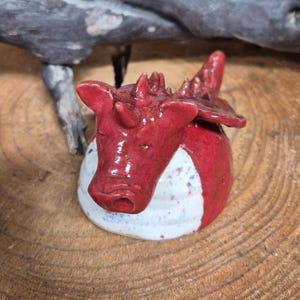May include: A handcrafted ceramic dragon ornament. The dragon is predominantly red with white and blue speckled detailing. The figurine has a rounded body and detailed features, including horns and wings. The dragon is resting on a wooden surface.